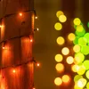 A wooden post wrapped with orange string lights, with blurred bokeh lights in yellow and green in the background. Náhled obrázku produktu