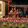 Rosa Solar-Blumenlichterkette, die einen grasbewachsenen Rasen vor einer Familie beleuchtet, die nachts auf einer Terrasse feiert Anteprima dell’immagine prodotto