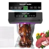 A vacuum sealer machine is shown sealing a cooked chicken in a plastic bag, with a stack of empty bags to the left and raw meat and vegetables to the right. The machine has a digital display and buttons. Náhľad obrázku produktu