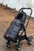 A black stroller with a black footmuff is parked outdoors on a path covered with fallen leaves, with a white structure in the background. Produktbild-Vorschau