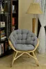 A rattan chair with a gray cushion sits in a room with a bookshelf and a lamp. Produktbild-Vorschau
