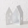 A white metal lantern shaped like a house, with cut-out windows and a hinged door, is displayed against a white brick wall. Produktbild-Vorschau