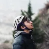 Mann mit Kletterhelm und Sonnenbrille schaut nach oben; trägt eine schwarze Jacke; Hintergrund mit unscharfen Bäumen. Podgląd zdjęcia produktu