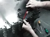 A person's hands are using a red tool to work on a large metal object, possibly a mold or machine part. Several screwdrivers are visible in the background. Produktbild-Vorschau