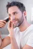 A man is trimming his beard in a bathroom mirror. Produktbild-Vorschau
