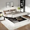 A modern bedroom with a white upholstered bed frame, lifted to reveal storage space underneath. The bed is made with white and brown linens and pillows. A bedside table with a lamp and a plant are visible to the left. A bookshelf is on the right. Produktbild-Vorschau