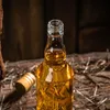 A close-up shot of a clear glass bottle filled with a golden liquid, placed on a wooden surface with some dry twigs scattered around. The bottle is open, and its cap lies nearby. Produktbild-Vorschau