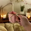A hand holds a pink, quilted mug with gold trim, set against a soft, fuzzy blanket and a softly lit background with candles and plants. Produktbild-Vorschau