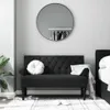 A dark gray tufted bench with a white round pillow and a white throw blanket draped over the armrest, placed in a room with a large round mirror on the wall and a bed on the right. Voir les images du produit
