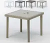 A square outdoor table with a woven rattan base and a glass top, shown in the foreground. Several smaller tables of the same design but in different colors (white, black, dark gray, light gray) are visible in the background. Anteprima dell’immagine prodotto