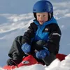 A child is sitting on a red sled in the snow, wearing a blue helmet and a blue and black jacket. Náhled obrázku produktu