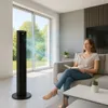 A woman is reading a book on a couch next to a tall black fan that is blowing air. Podgląd zdjęcia produktu