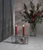 Two red LED taper candles in glass candle holders with a dried rose inside one, placed on a white wooden table with a bouquet of flowers in a vase in the background. Produktbild-Vorschau