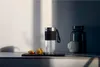 A glass bottle with a black grip and silver lid sits on a dark tray, next to two physalis fruits. In the background, a dark blue bottle and a light gray mug are visible. Voir les images du produit