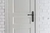 A close-up view of a white door with a black door handle, set against a white brick wall with an electrical outlet. Produktbild-Vorschau