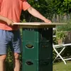 A person is assembling a table outdoors using green plastic crates as a base and a wooden board as the tabletop. There is a white chair and plants in the background. Produktbild-Vorschau