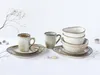 A set of ceramic tableware, including mugs and bowls, arranged on a white surface with decorative dried branches and stones. Produktbild-Vorschau