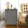 A gray nightstand with a drawer and a door sits next to a cream-colored armchair with a decorative pillow. A lamp, a wooden hand sculpture, and a cup of coffee are on top of the nightstand. Náhľad obrázku produktu