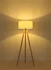 A floor lamp with a fabric shade and wooden tripod legs, illuminated against a plain wall. Produktbild-Vorschau