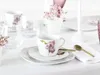A close-up of a white porcelain teacup and saucer with a purple orchid design, set on a white tablecloth with other tableware and floral decorations. Produktbild-Vorschau