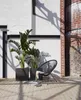 Two large dark gray square planters with lush green plants are placed on a paved patio next to a black woven chair. A brick building is in the background. Produktbild-Vorschau