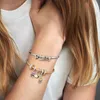 A close-up shot of a person's arm wearing two Pandora charm bracelets, one silver with letter charms spelling 'PLAY' and another gold with various charms. Podgląd zdjęcia produktu