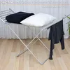 A silver drying rack with a black blanket and a white pillow on top, and a dark gray shirt hanging off the side. The background is a white curtain and a light wood floor. Produktbild-Vorschau