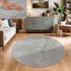 A circular gray rug is placed on a wooden floor in a living room setting, with a sofa, cabinet, and plant visible. Voir les images du produit