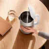 A hand is holding a white electric kettle on a wooden table, with a glass of water and a book nearby. Náhľad obrázku produktu