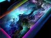A close-up, angled view of a gaming mousepad with a colorful, abstract cloud-like design, illuminated by RGB lighting around the edges. Náhľad obrázku produktu