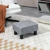 A gray upholstered ottoman sits on a patterned rug in a living room setting, with a white chair and a brown throw blanket visible. Produktbild-Vorschau