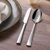 A close-up shot of a knife and spoon set on a wooden table, next to a plate with a napkin. Produktbild-Vorschau