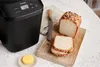A black Panasonic bread maker is shown next to a loaf of bread that has been sliced on a wooden cutting board. A pat of butter is in a small dish. Náhľad obrázku produktu