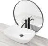 A white oval countertop sink with black faucets is shown on a light wood surface, with a round black-framed mirror reflecting the scene. Náhled obrázku produktu