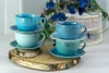 Four ceramic cups and saucers are stacked in pairs on a wooden base, with blue flowers in the background. Náhľad obrázku produktu