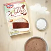 A flat lay of a Dr. Oetker Schoko Wolke cake mix box, a slice of chocolate cake, a small bowl of powdered sugar, and a bowl of cake mix powder, all arranged on a wooden surface. Produktbild-Vorschau