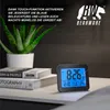 A digital alarm clock is placed on a nightstand next to books and a plant, with promotional text and a brand logo in the background. Podgląd zdjęcia produktu