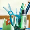 A close-up of a glass cup filled with various writing utensils, including scissors, pens, and markers. A green paper crown with an orange flower is visible in the foreground. Náhled obrázku produktu