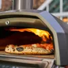 A pizza is baking in an outdoor pizza oven with flames visible. Produktbild-Vorschau