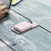 A pink power bank is placed on top of a silver smartphone, which is resting on a wooden surface. A person's hand is visible in the background, interacting with a laptop. Náhled obrázku produktu