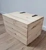 A wooden storage box with a lid sits on a wooden floor in front of a light gray wall. Náhled obrázku produktu