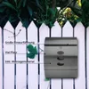 A stainless steel mailbox mounted on a white picket fence, with green foliage in the background. Produktbild-Vorschau