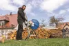 A man is using a log saw in a yard with piles of firewood in the background. Náhľad obrázku produktu