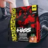 A bag of protein powder is placed in front of a barbell and weight plates, with a person's legs and feet visible in the background, suggesting a gym environment. Produktbild-Vorschau