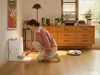 A woman is kneeling on the floor, interacting with a white robot vacuum cleaner and a charging station. The scene is set in a well-lit room with wooden floors and furniture. Produktbild-Vorschau