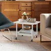 A white rolling side table with items on it, placed in a living room setting next to a chair and a couch, with a wooden cabinet in the background. Produktbild-Vorschau