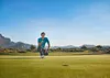 A man is crouching on a golf course, holding a golf putter, with mountains in the background. Produktbild-Vorschau