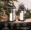 Two black lanterns with LED candles inside, placed on a wooden surface with plants in the background. Podgląd zdjęcia produktu