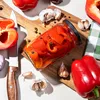 A close-up overhead view of pickled red peppers in a glass jar, surrounded by fresh red bell peppers, garlic, and peppercorns on a wooden cutting board. Podgląd zdjęcia produktu