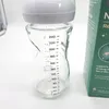 A close-up of a glass baby bottle with measurement markings and a white cap, next to a green product box. Podgląd zdjęcia produktu